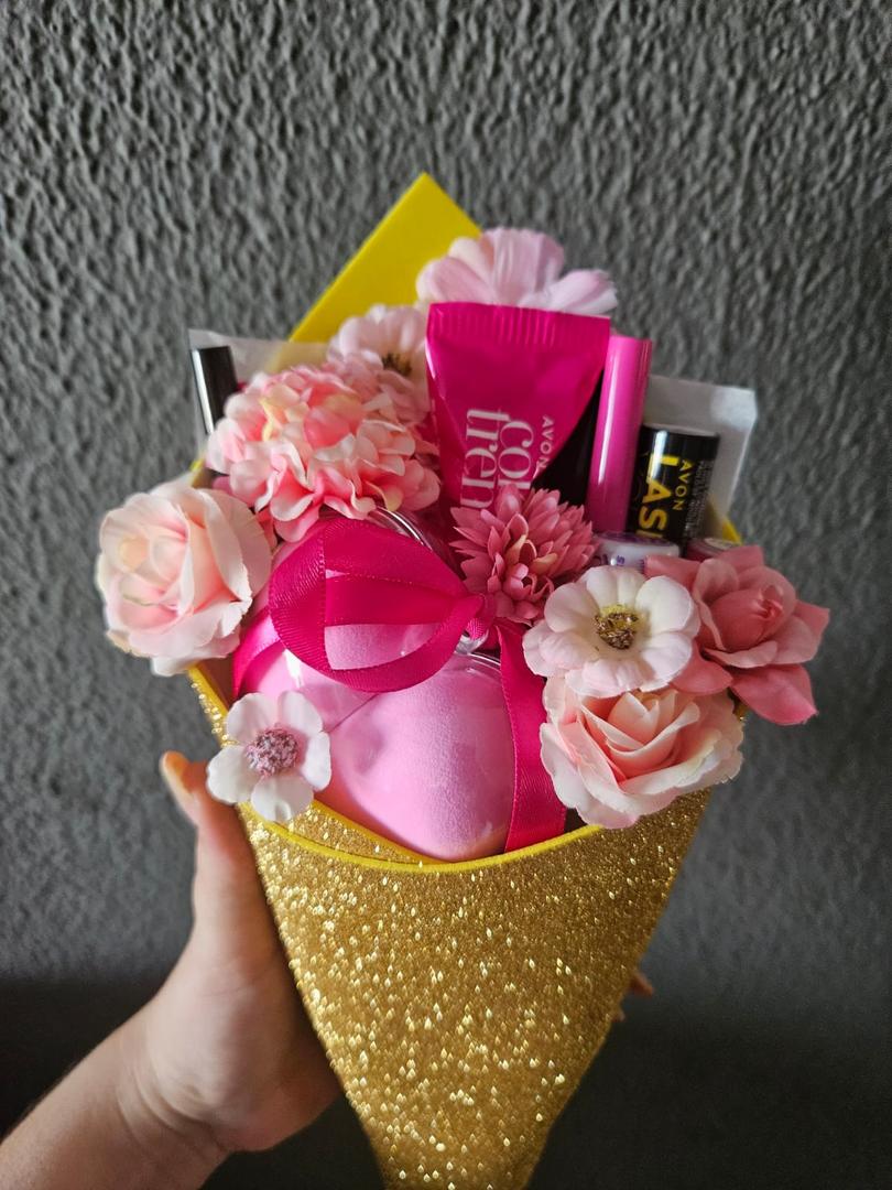 Na imagem temos um Buque feito com flores e maquiagens Avon. As flores são em tons de rosa, de maquiagens temos diversos produtos como lápis de olho, delineador, esponja de maquiagem, máscara de cílios, paleta de sombras, batom e blush, e gloss labial. Está envonvido com uma folha de E.V.A amarelo com glitter. E aparece a mão de uma pessoa segurando o Buque.